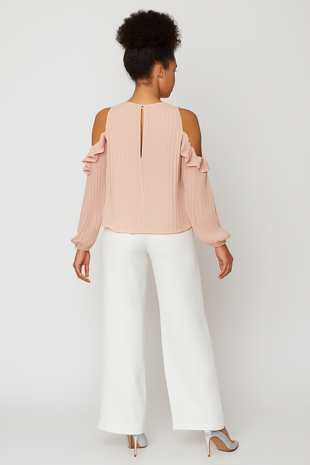Woman wearing a light pink blouse with cold shoulder design and white pants on a plain background