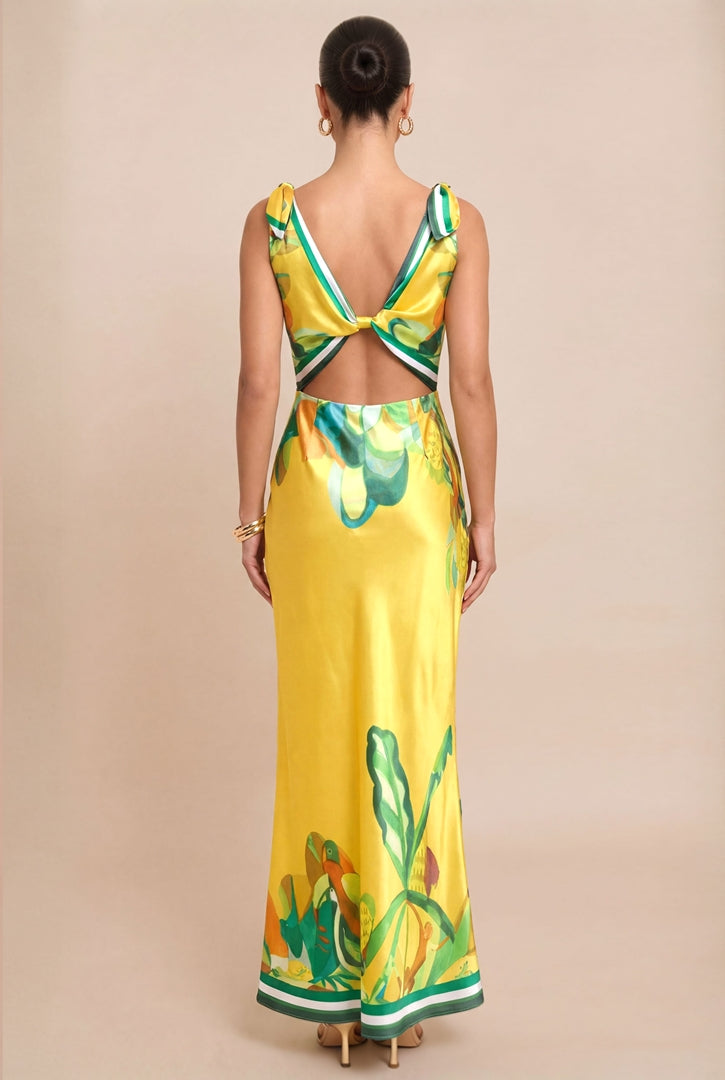 A woman viewed from the back wearing a yellow satin maxi dress with green and white border details and a green floral print. The dress features a deep V-back with a bow detail at the center.