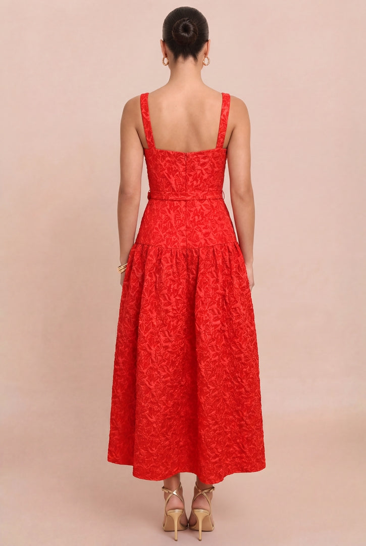 A woman viewed from the back wearing a bright red sleeveless midi dress with a textured floral lace pattern, a fitted bodice, and a flared skirt, paired with beige high-heeled sandals against a solid beige studio background.