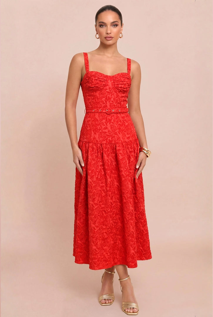A woman modeling a bright red midi dress with a textured floral lace pattern, featuring a ruched sweetheart bodice, thin shoulder straps, and a flared skirt, set against a solid pale pink background.