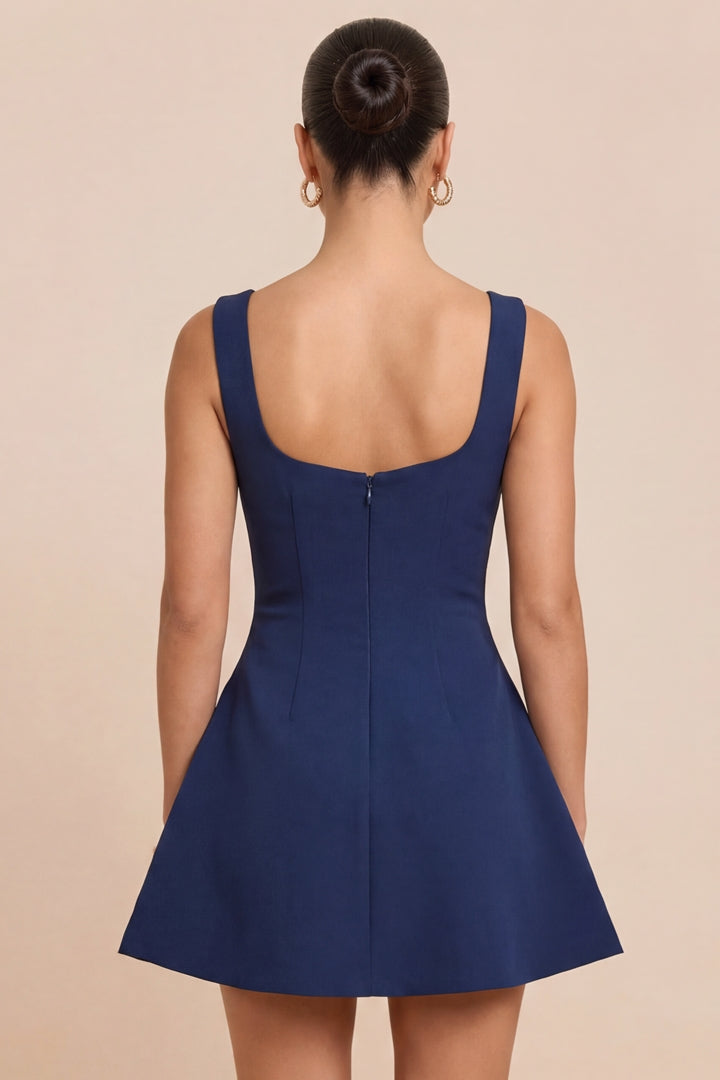 A woman shown from the back wearing a navy blue sleeveless fit-and-flare dress with a square neckline and a zip closure at the back, set against a solid beige background.