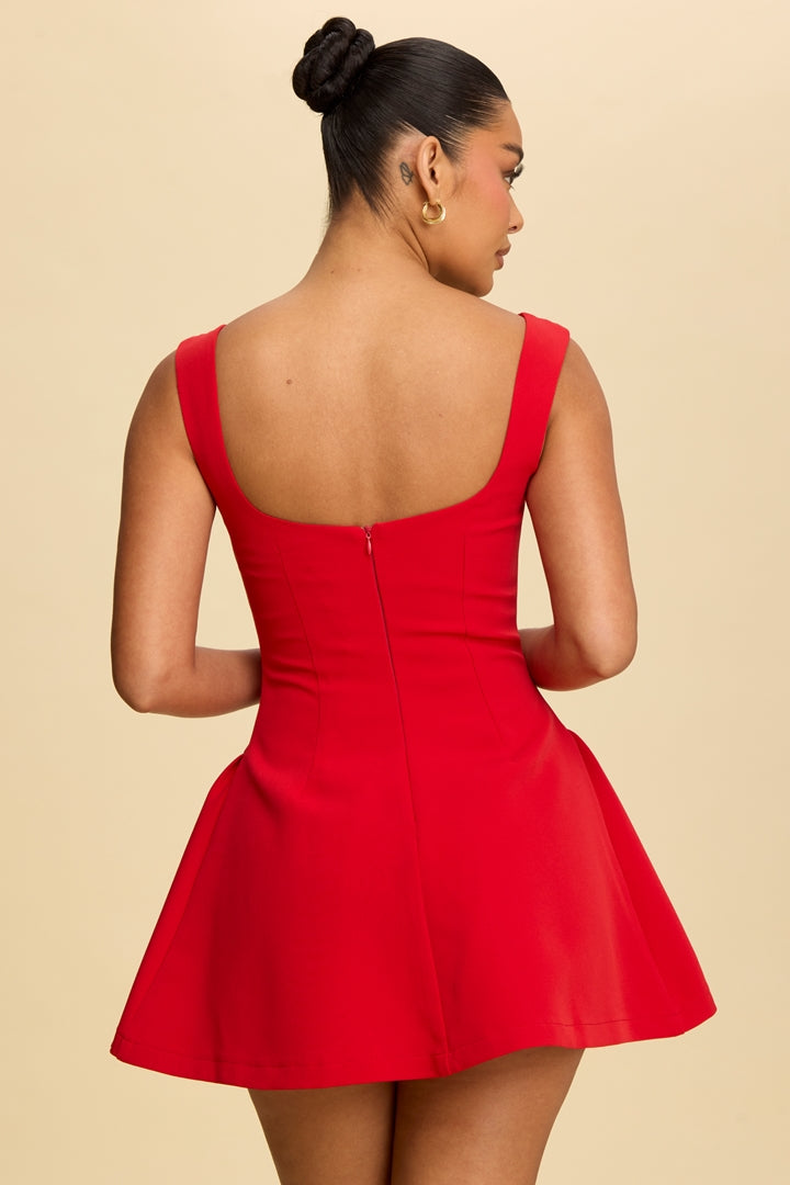 A woman shown from the back wearing a bright red sleeveless fit-and-flare dress with a scoop open back, photographed against a plain warm beige studio background.