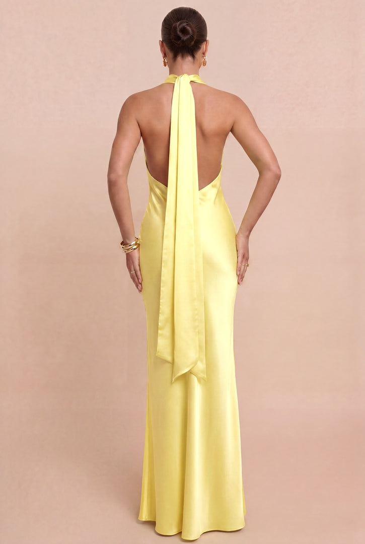 A woman viewed from the back wearing a floor-length satin gown in a pale pastel yellow. The dress has an open back with a long draped tie and a fitted silhouette, shown against a plain warm beige studio background.