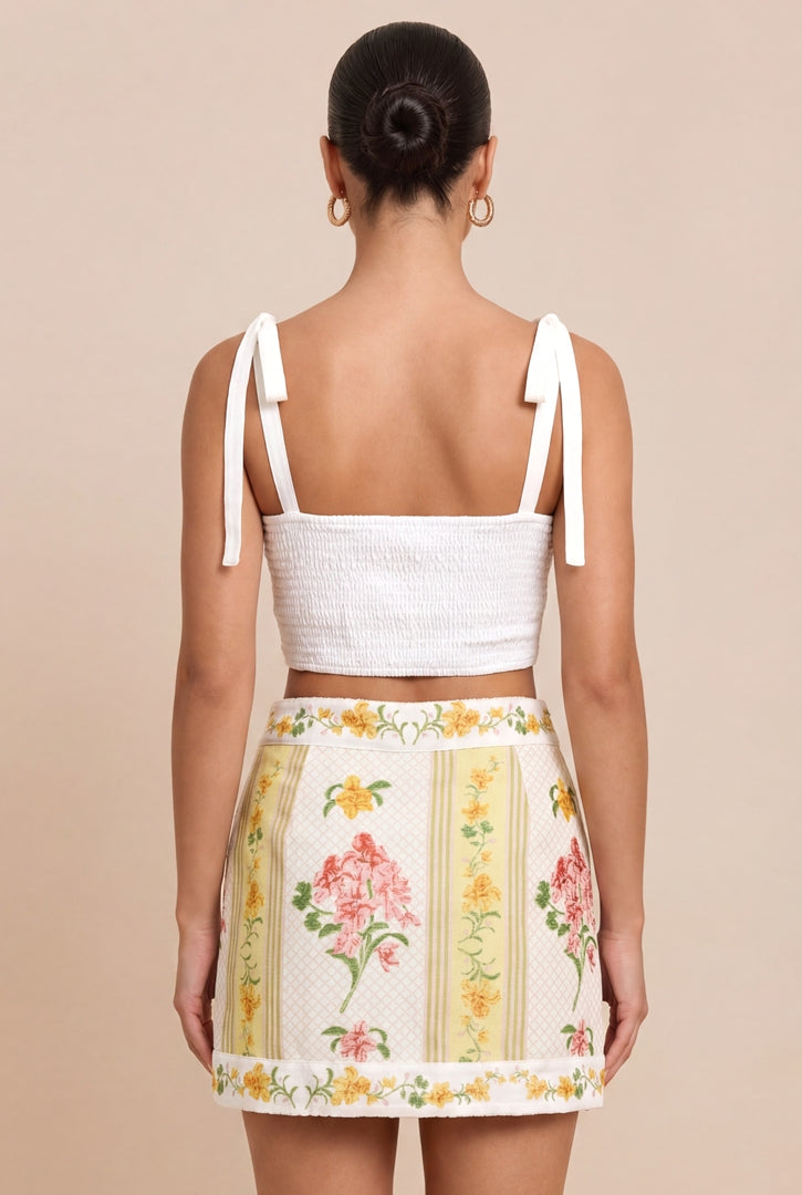 A woman shown from the back wearing a white crop top with tie straps and a cream mini skirt with a yellow, pink, and green floral print, set against a solid beige background.