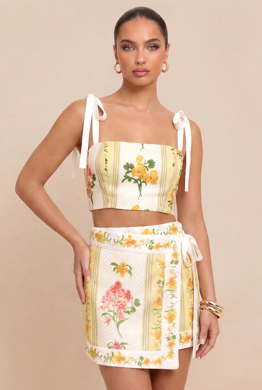 A model wearing a two-piece matching set consisting of a cream-colored crop top with yellow floral prints and tie straps, and a matching wrap-style mini skirt with pink floral prints and yellow borders, set against a solid beige background.
